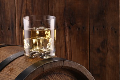 Whiskey with ice cubes in glass on barrel against wooden background, space for text