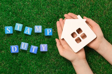 Woman holding house figure and blue wooden cubes with text Stamp Duty on green grass, top view