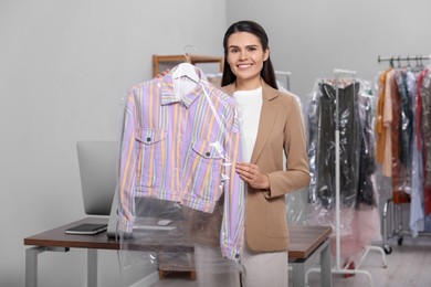 Photo of Dry-cleaning service. Happy woman holding hanger with shirt in plastic bag indoors