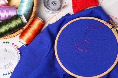 Blue cloth with hoop, embroidered heart, thread and sewing needle on light table with tools, flat lay