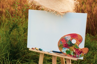 Photo of Wooden easel with blank canvas, painting equipment and hat in field. Space for text