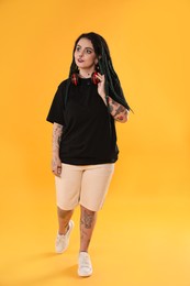 Photo of Beautiful young woman with tattoos on body, nose piercing and dreadlocks against yellow background
