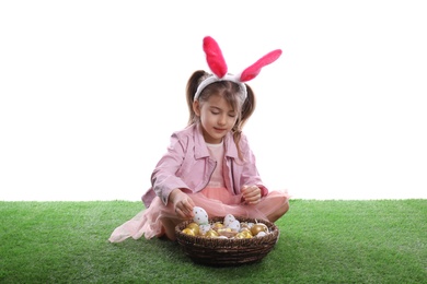 Photo of Adorable little girl with bunny ears and wicker basket full of Easter eggs on green grass against white background
