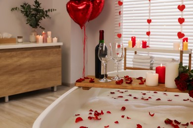 Photo of Wooden tray with wine, burning candles and rose petals on tub in bathroom. Valentine's day celebration