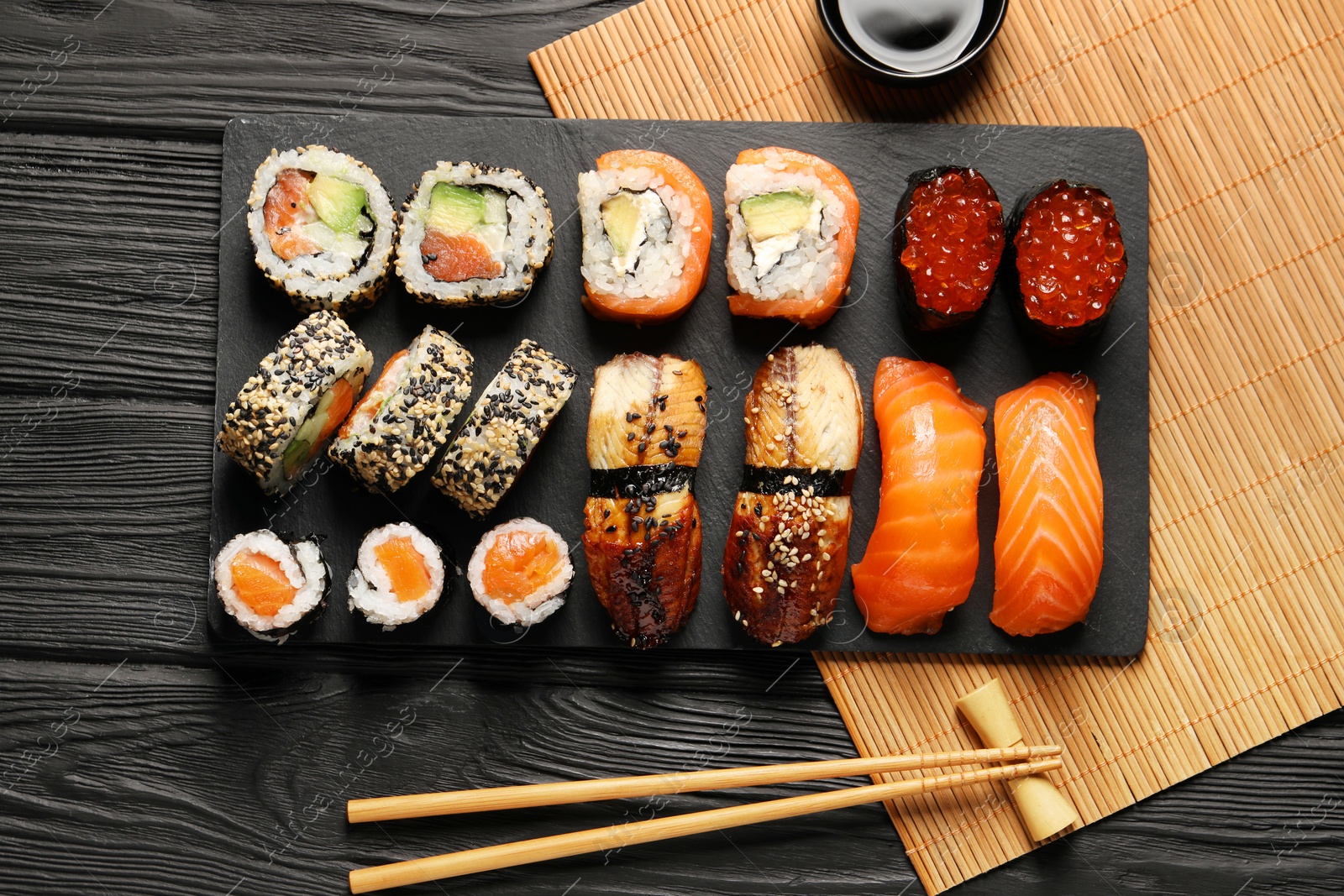 Photo of Delicious sushi rolls, soy sauce and chopsticks on black wooden table, flat lay