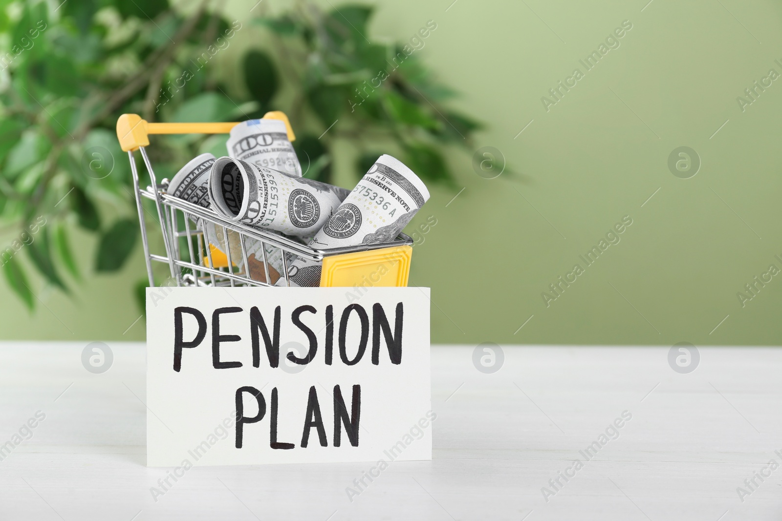 Photo of Card with phrase Pension Plan and dollar banknotes in toy shopping cart on white wooden table, space for text. Retirement concept