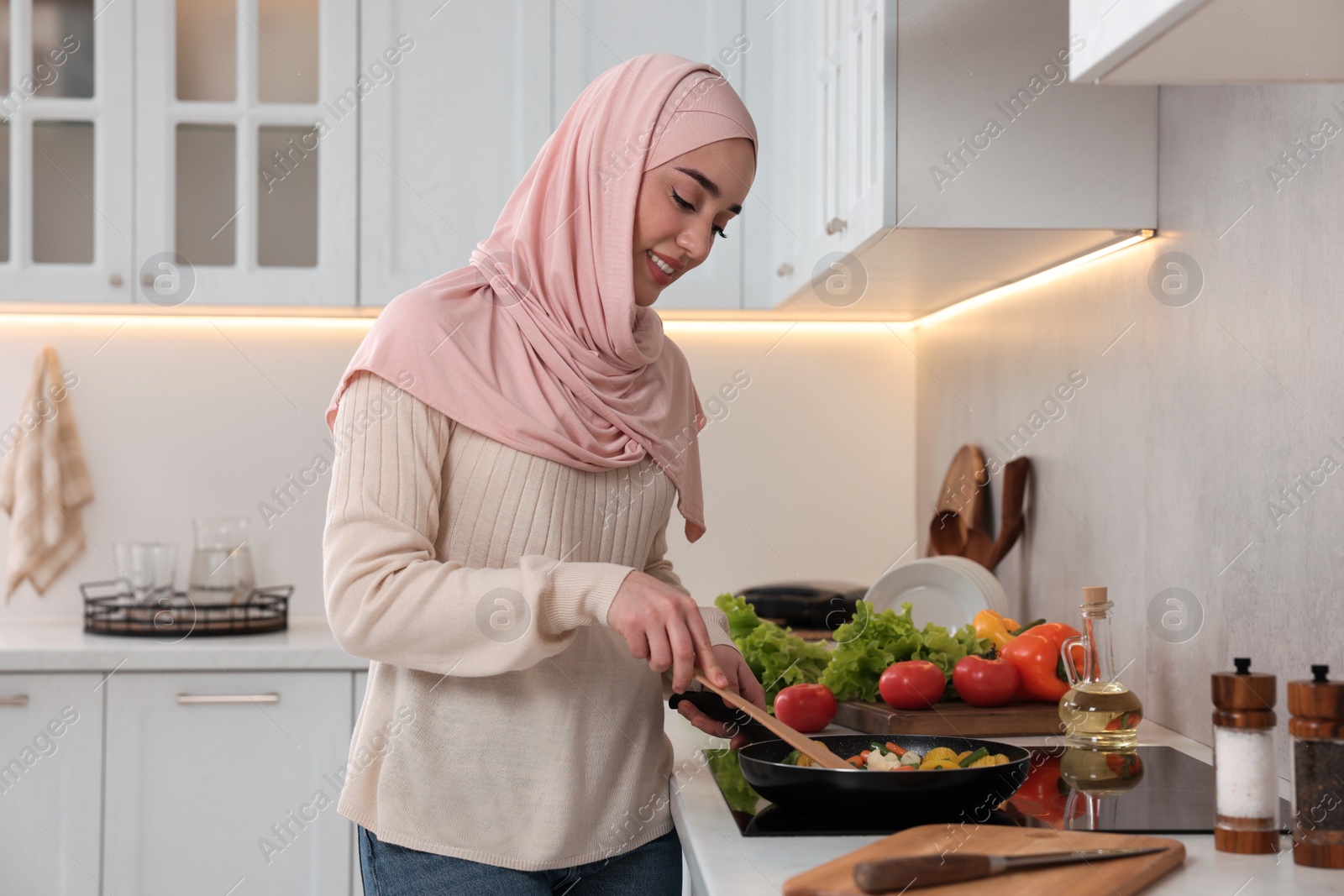 Photo of Muslim woman cooking delicious dish with vegetables on cooktop in kitchen