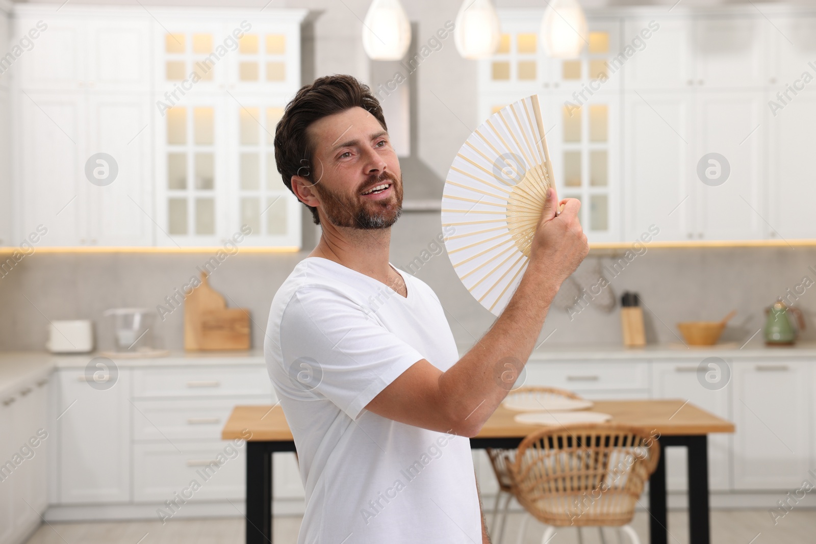 Photo of Bearded man waving white hand fan to cool himself in kitchen