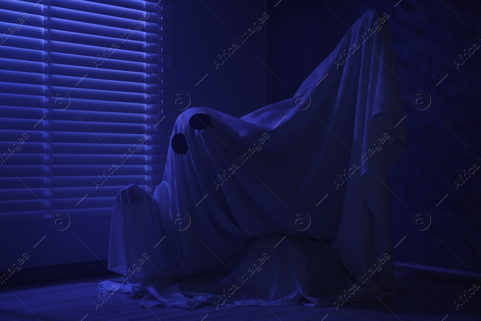 Photo of Creepy ghost. Woman covered with sheet near window in blue light