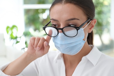 Photo of Woman wiping foggy glasses caused by wearing medical mask indoors, closeup