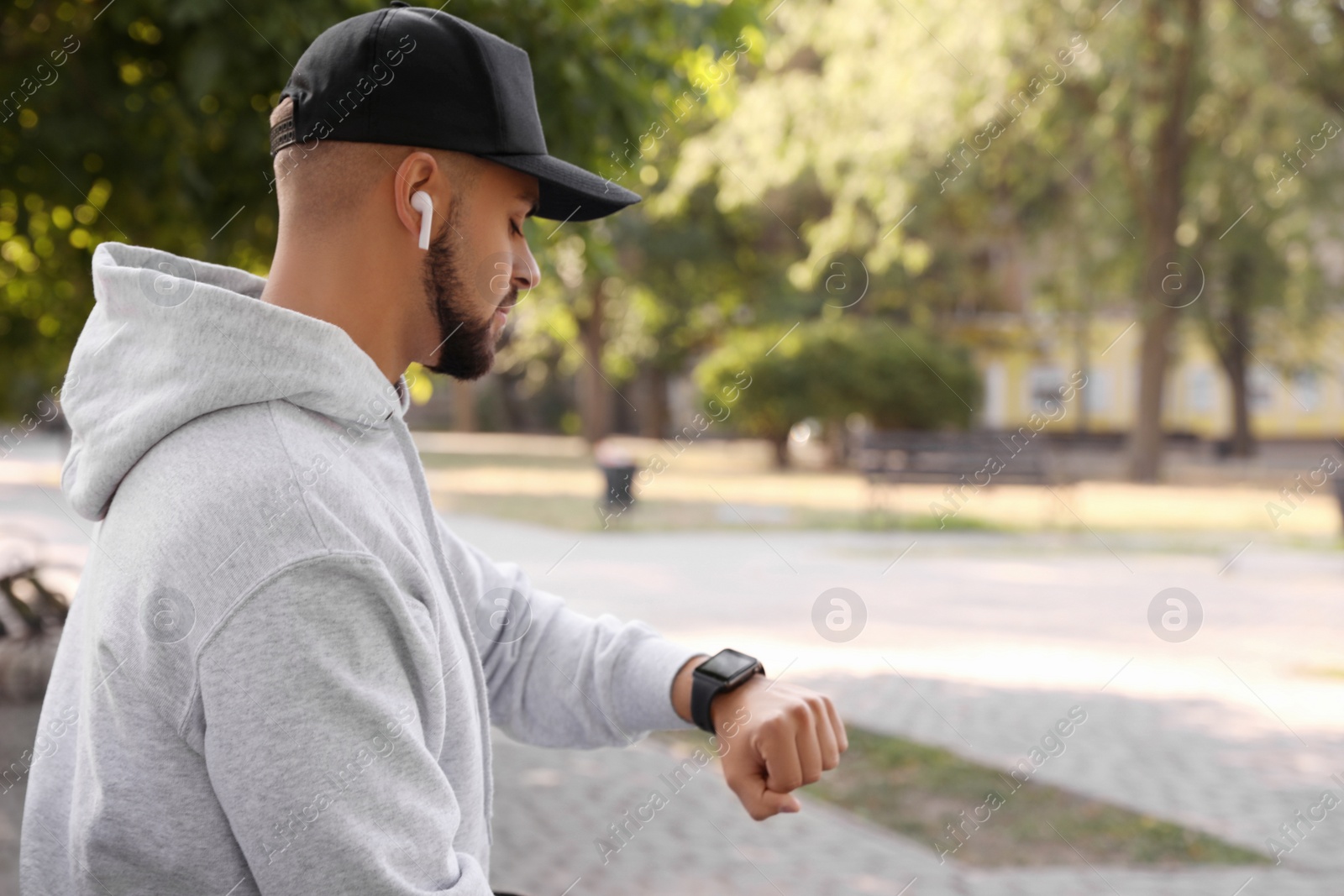 Photo of Young man with wireless headphones and smartwatch listening to music in park. Space for text