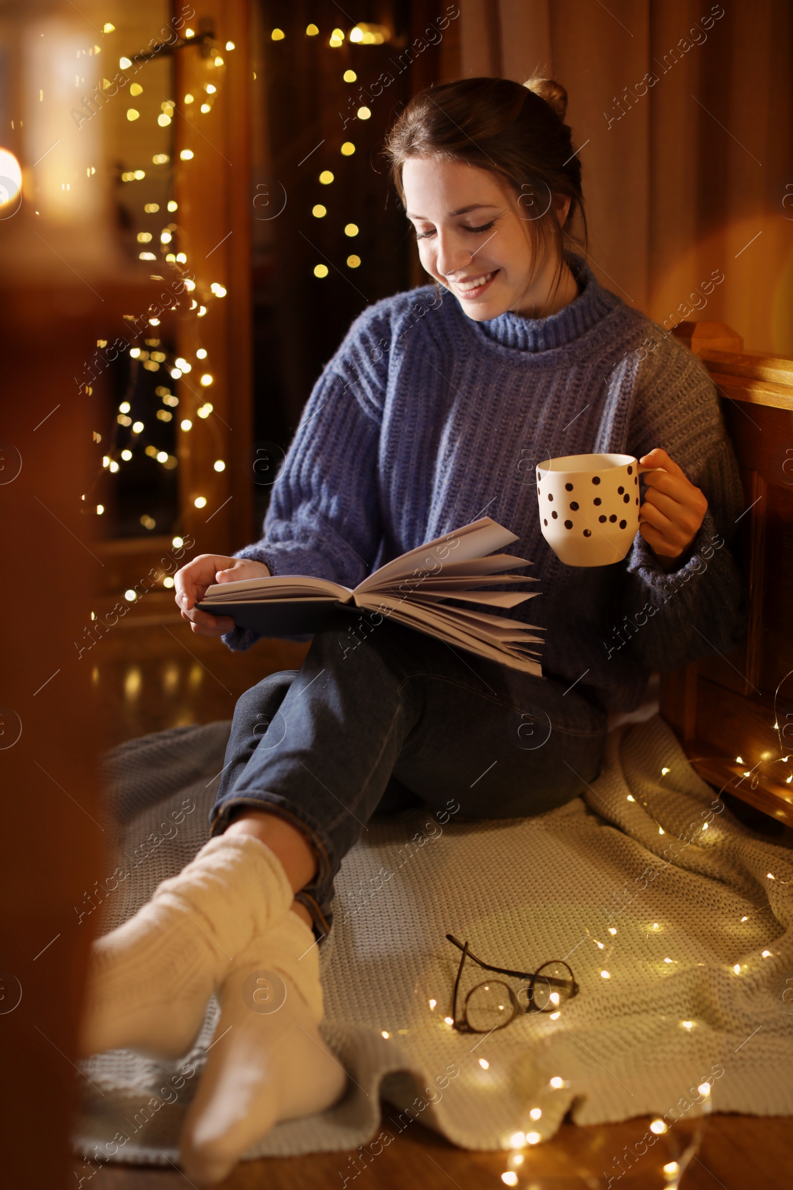 Photo of Woman with cup of hot beverage reading book at home in winter evening