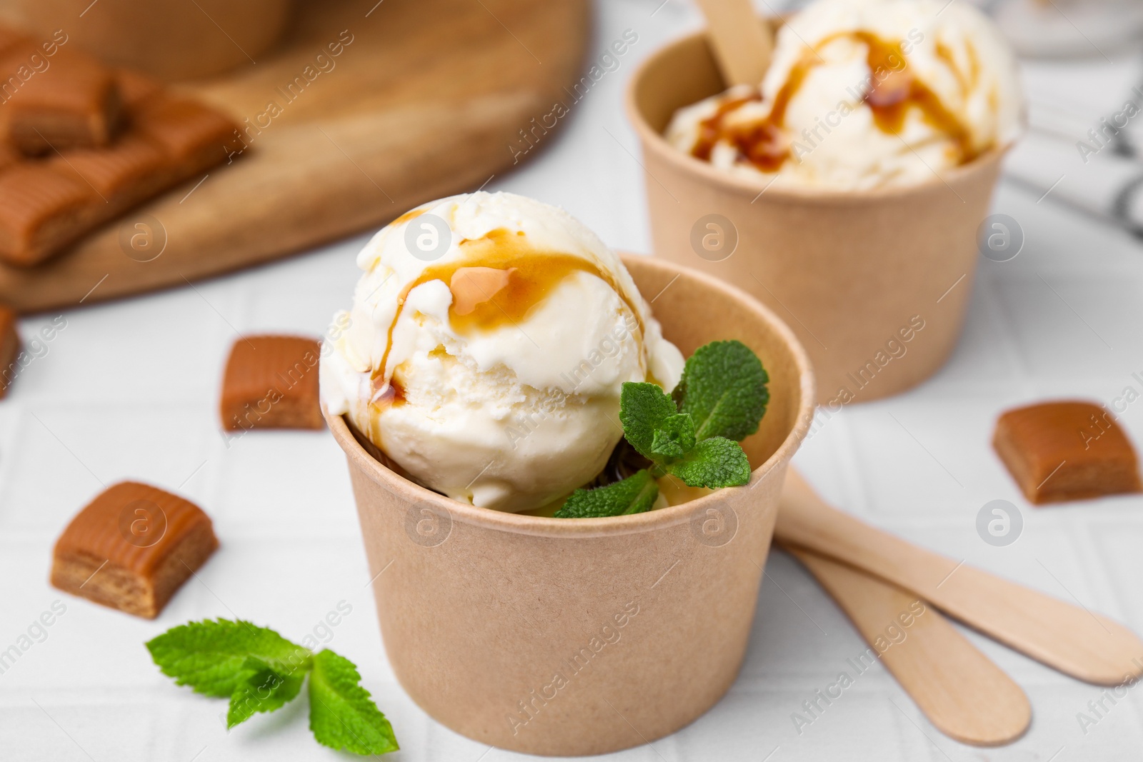 Photo of Scoops of ice cream with caramel sauce, mint leaves and candies on white tiled table, closeup