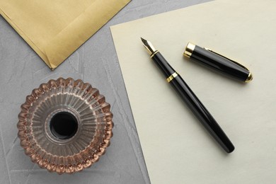 Stylish fountain pen, inkwell, envelope and paper on grey textured table, flat lay