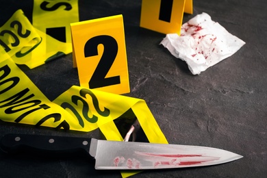 Photo of Bloody knife, yellow tape and evidence marker on black slate table, closeup. Crime scene