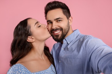Photo of Woman kissing her smiling boyfriend while he taking selfie on pink background
