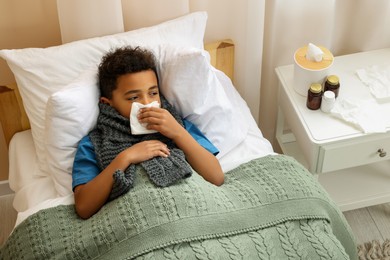 Photo of African-American boy with scarf and tissue blowing nose in bed indoors, above view. Cold symptoms