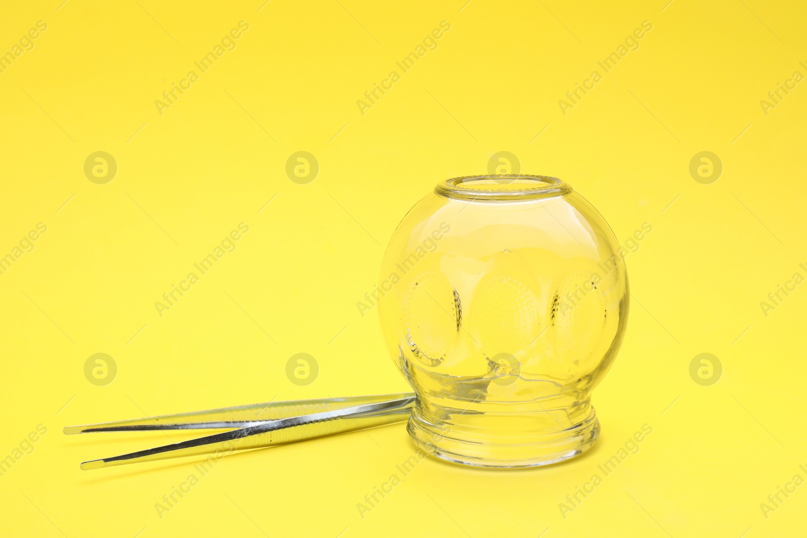 Photo of Glass cup and tweezers on yellow background, closeup. Cupping therapy