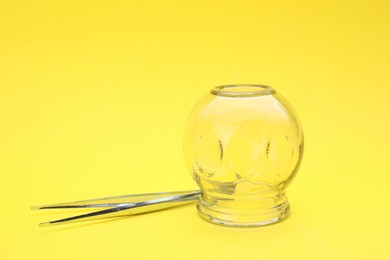 Photo of Glass cup and tweezers on yellow background, closeup. Cupping therapy