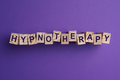 Photo of Wooden blocks with word HYPNOTHERAPY on purple background, flat lay