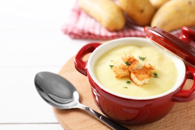 Photo of Tasty potato soup with croutons in ceramic pot and spoon on white wooden table