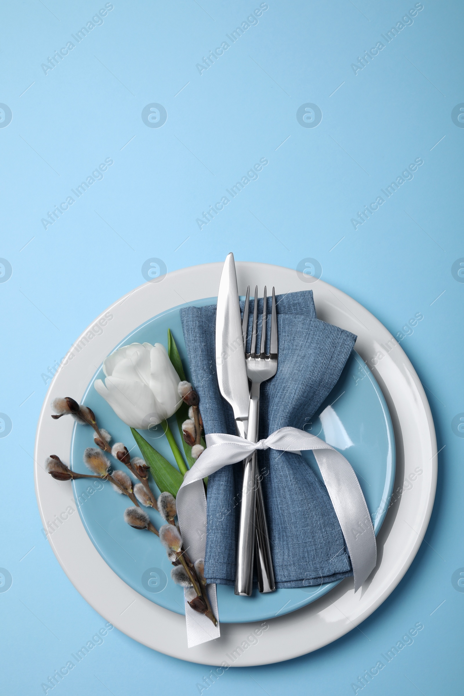 Photo of Festive table setting with willow twigs and tulip on light blue background, top view. Easter celebration