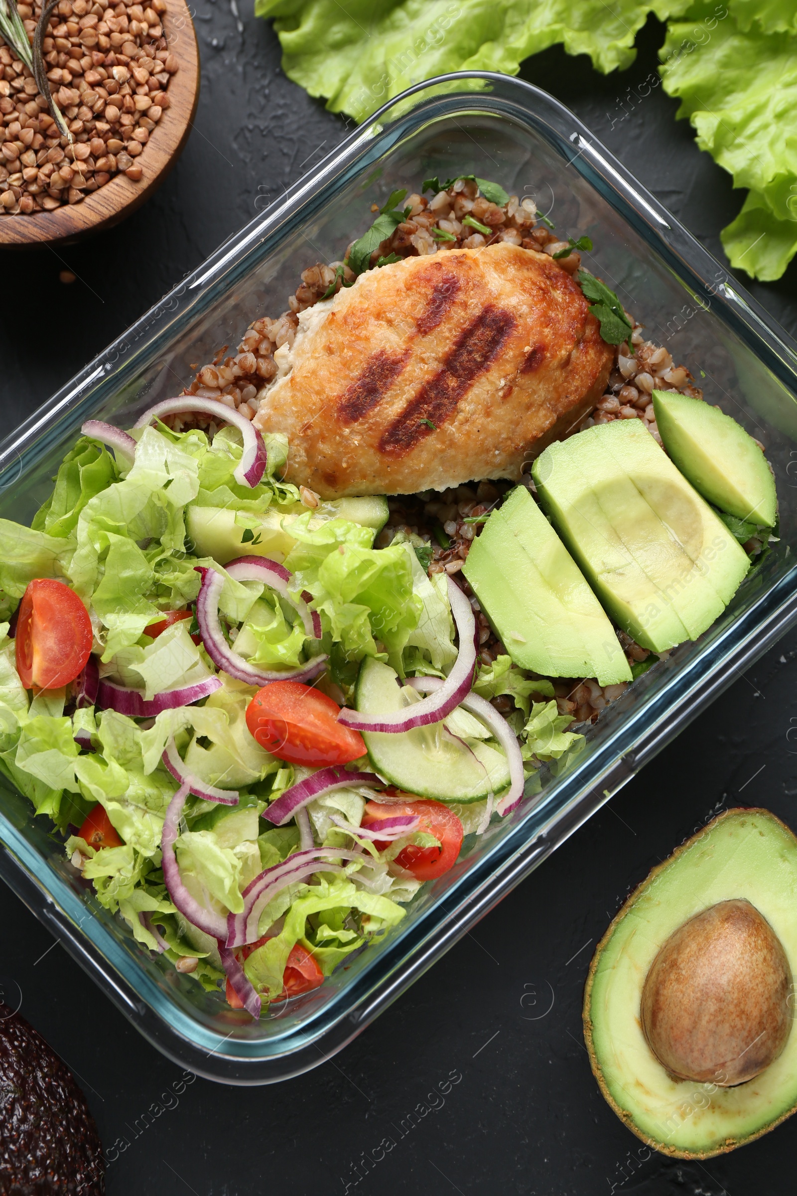 Photo of Healthy meal. Fresh salad, avocado, cutlet and buckwheat in glass container near other products on black table, flat lay