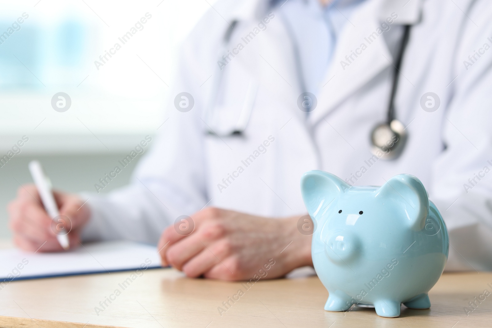 Photo of Doctor writing notes at wooden table, focus on piggy bank