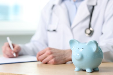 Photo of Doctor writing notes at wooden table, focus on piggy bank