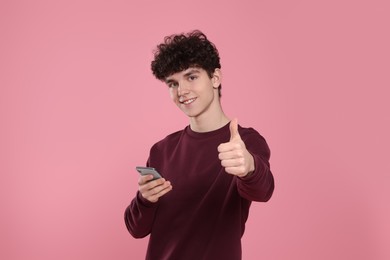 Photo of Portrait of smiling teenage boy with smartphone showing thumb up on pink background