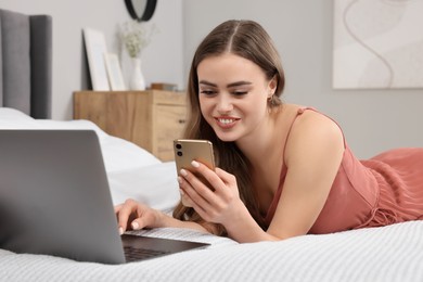 Photo of Happy woman with smartphone and laptop on bed in bedroom