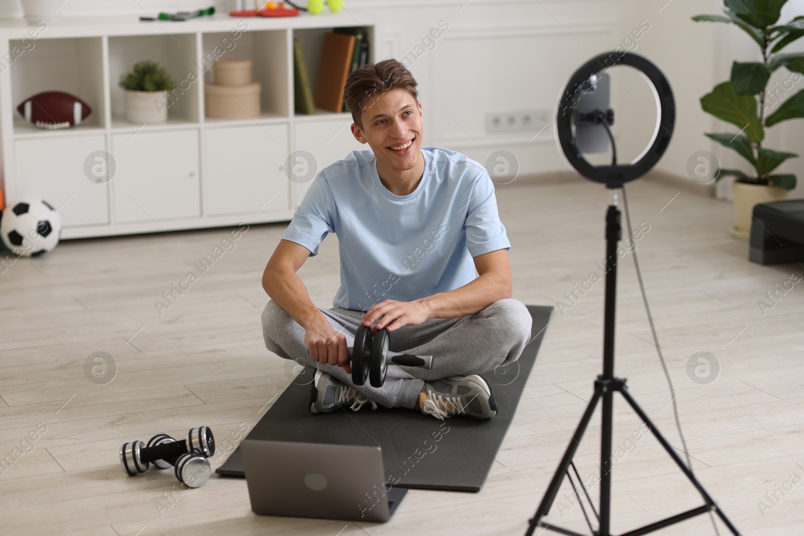 Photo of Smiling sports blogger holding ab roller while streaming online fitness lesson with smartphone at home