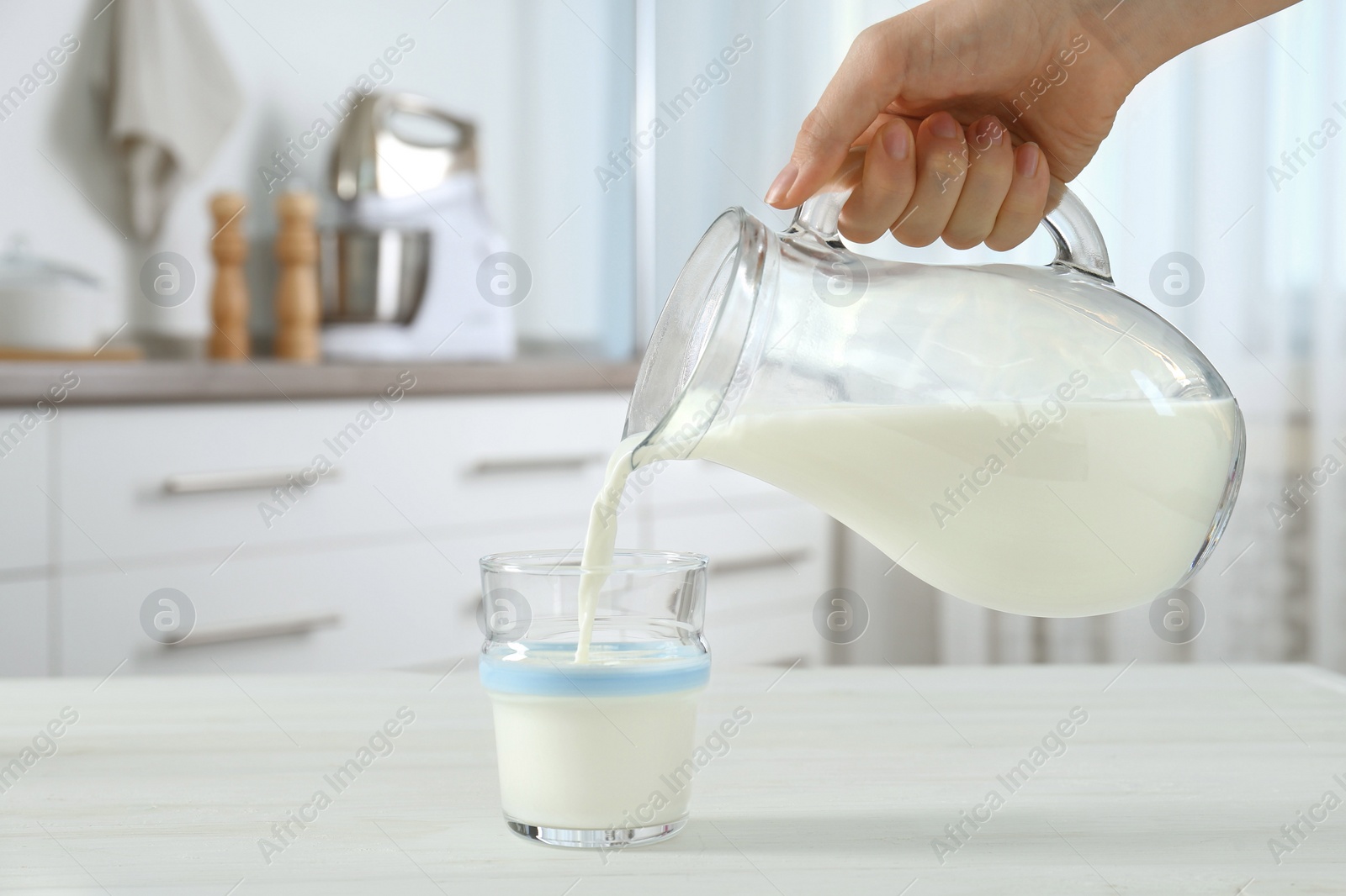 Photo of Woman pouring milk from jug into glass at white wooden table in kitchen, closeup
