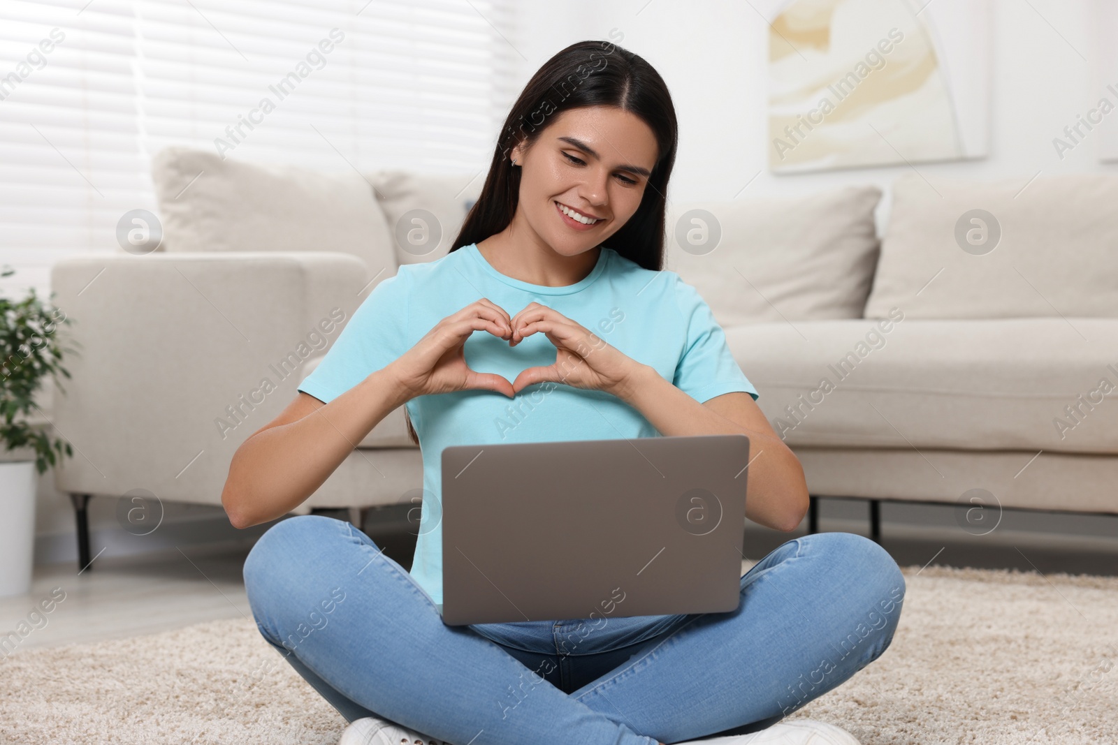 Photo of Happy young woman having video chat via laptop and making heart on floor in living room