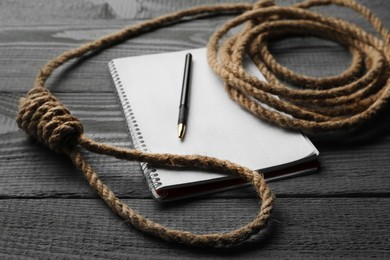 Photo of Rope noose and blank notebook with pen on grey wooden table, closeup