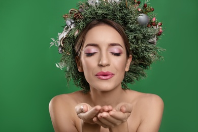 Beautiful young woman with Christmas wreath blowing kiss on green background