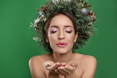 Photo of Beautiful young woman with Christmas wreath blowing kiss on green background