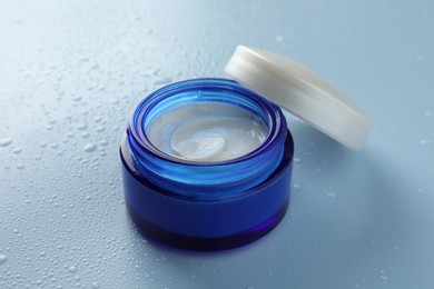 Moisturizing cream in open jar on light blue background with water drops, closeup