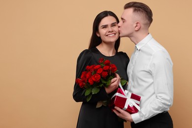 Photo of Lovely couple with gift box and bouquet of red roses on beige background, space for text. Valentine's day celebration