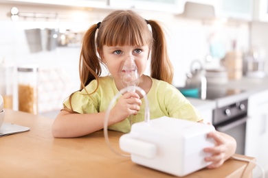 Photo of Little girl using asthma machine at table in kitchen