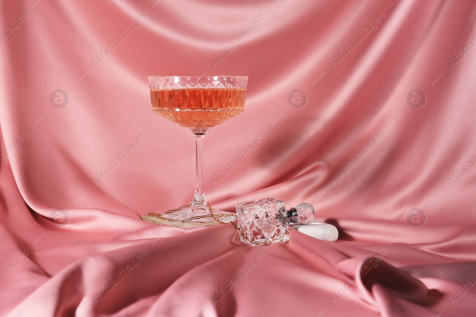 Photo of Glass of alcohol drink, perfume and jewelry on pink cloth