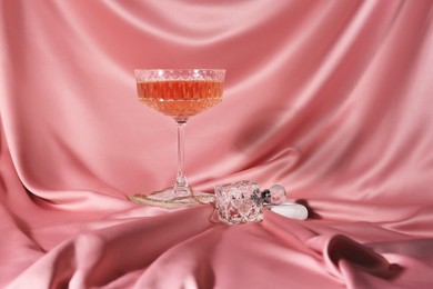 Glass of alcohol drink, perfume and jewelry on pink cloth