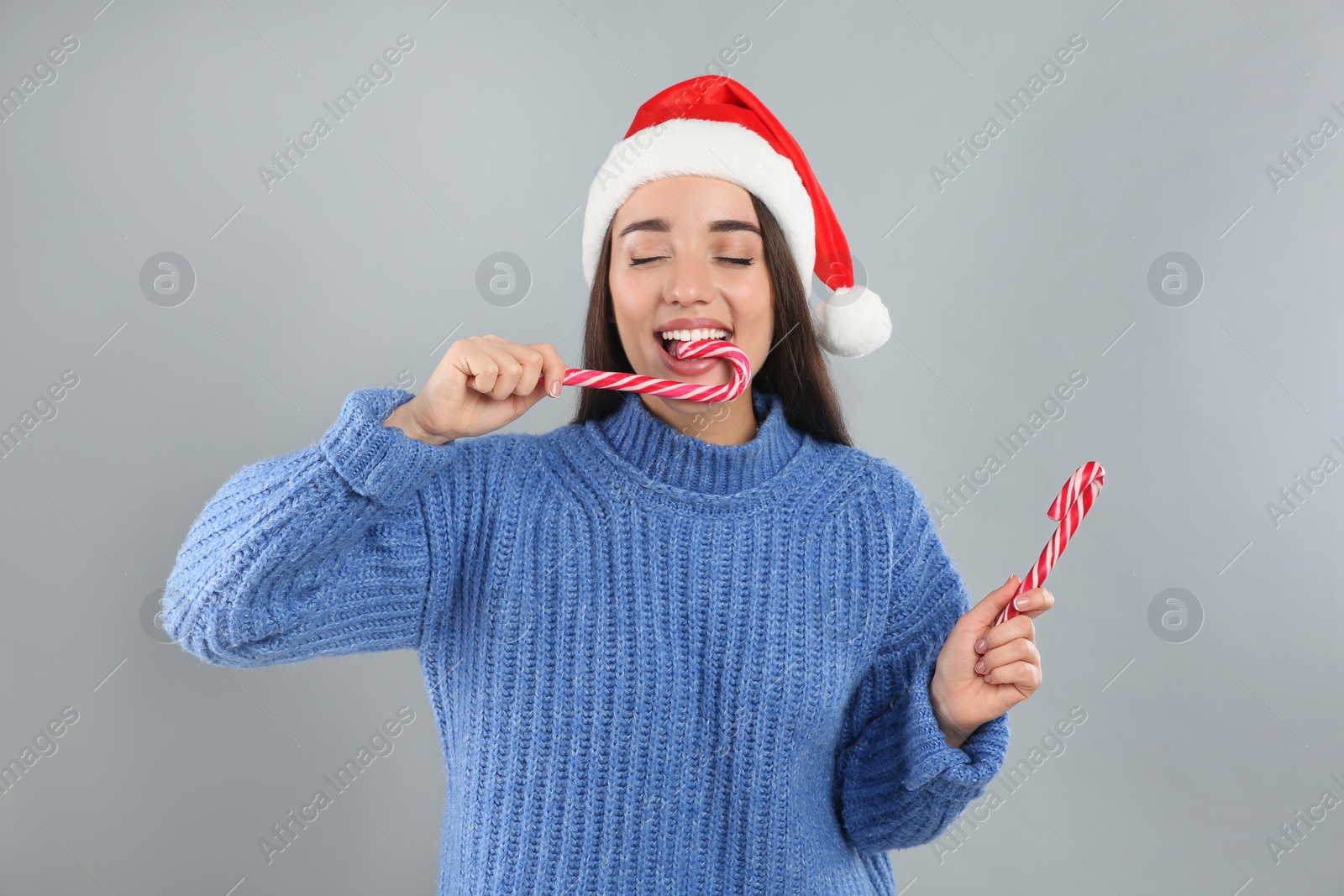 Photo of Young woman in blue sweater and Santa hat eating candy canes on grey background. Celebrating Christmas