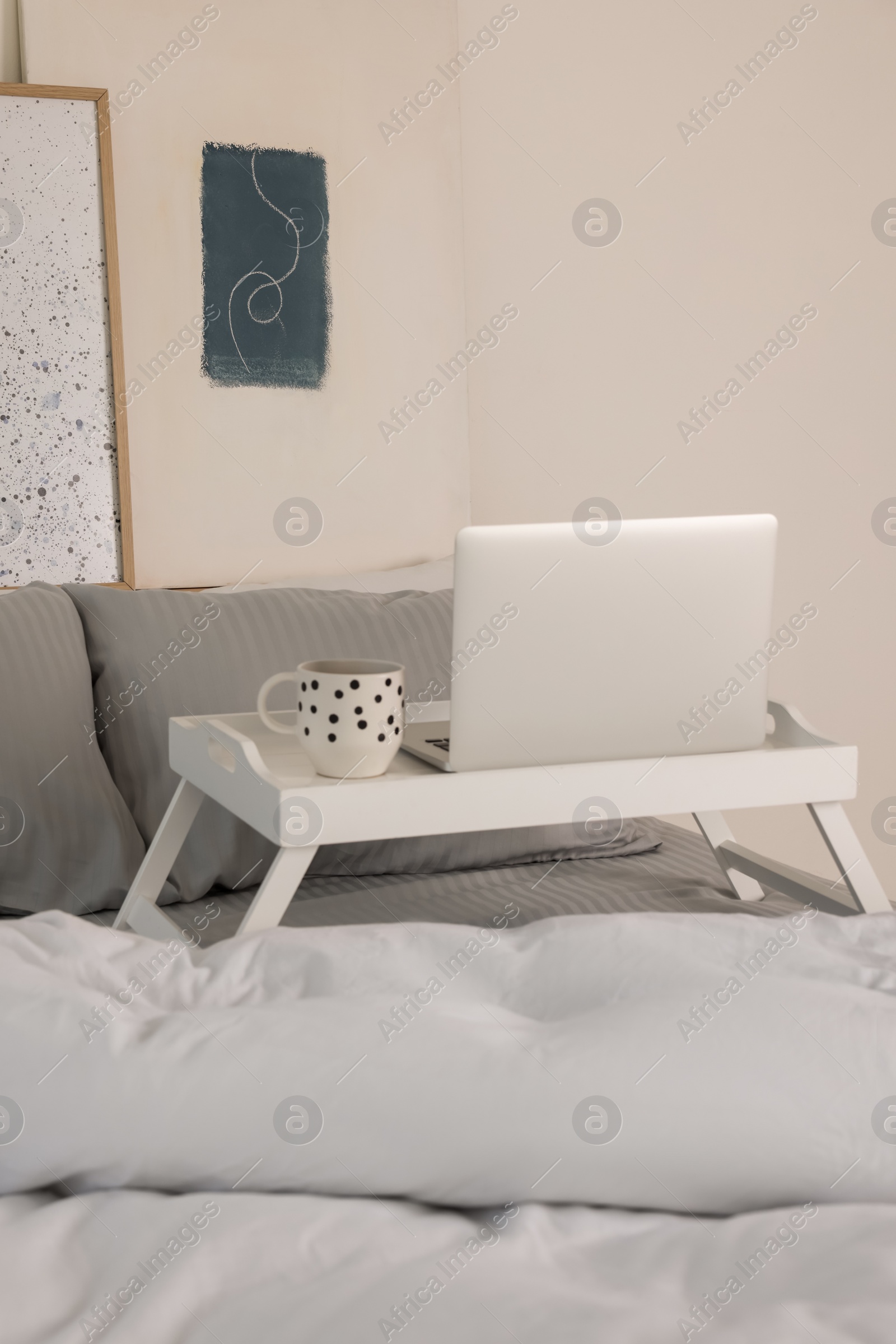 Photo of White tray table with laptop and cup of drink on bed indoors