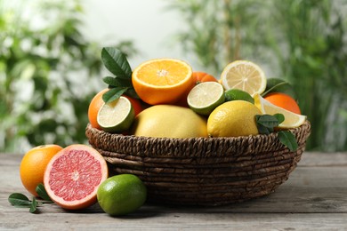 Different fresh citrus fruits and leaves in wicker basket on wooden table against blurred background, closeup