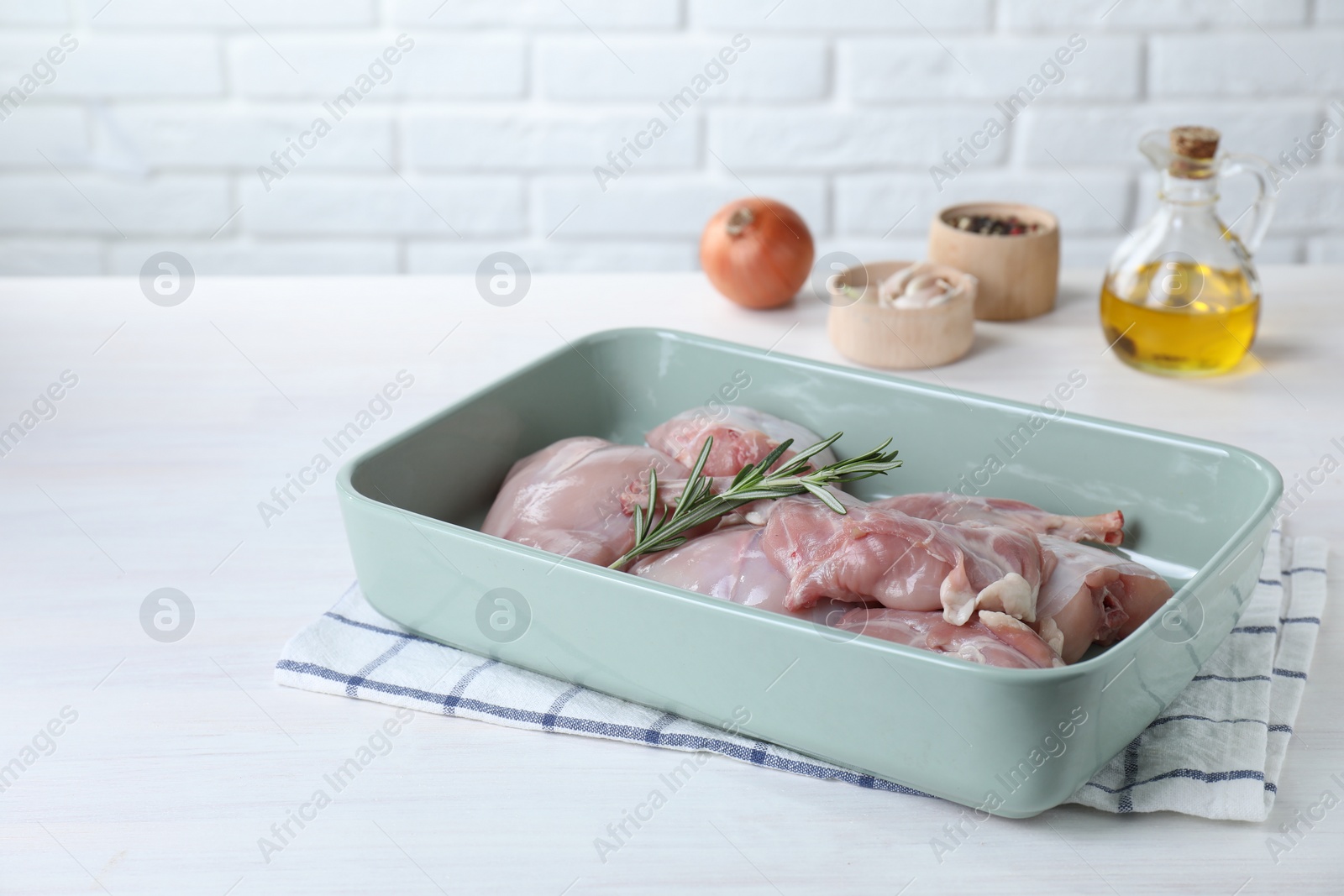 Photo of Raw rabbit meat and rosemary in baking dish on white wooden table, space for text