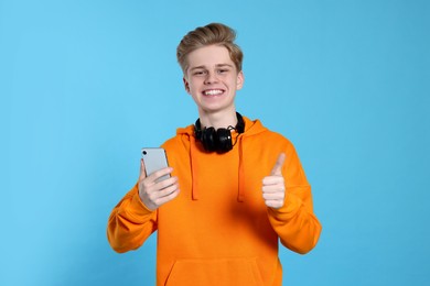 Photo of Teenage boy with smartphone and headphones showing thumb up on light blue background