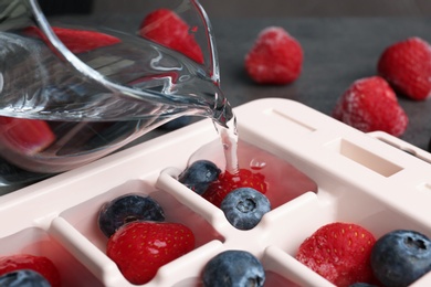 Pouring water into ice cube tray with berries on table, closeup