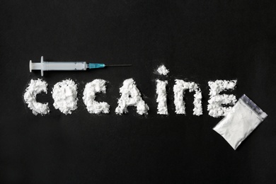 Photo of Word Cocaine written with white powder, full plastic bag and syringe on black background, top view