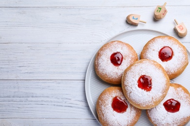 Photo of Hanukkah doughnuts with jelly and sugar powder near dreidels on white wooden table, flat lay. Space for text.